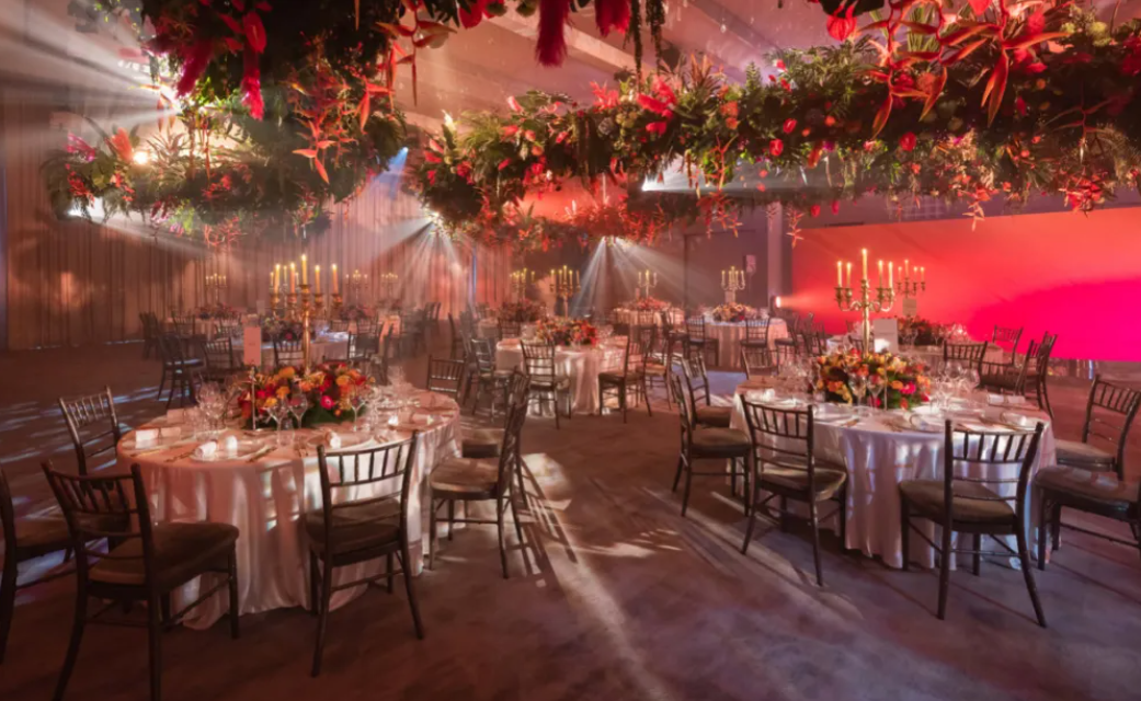 An indoor high jewelry dinner at the Eden: Garden of Wonders event in Bangkok, with candlelit round banquet tables beneath suspended floral installations and beams of light