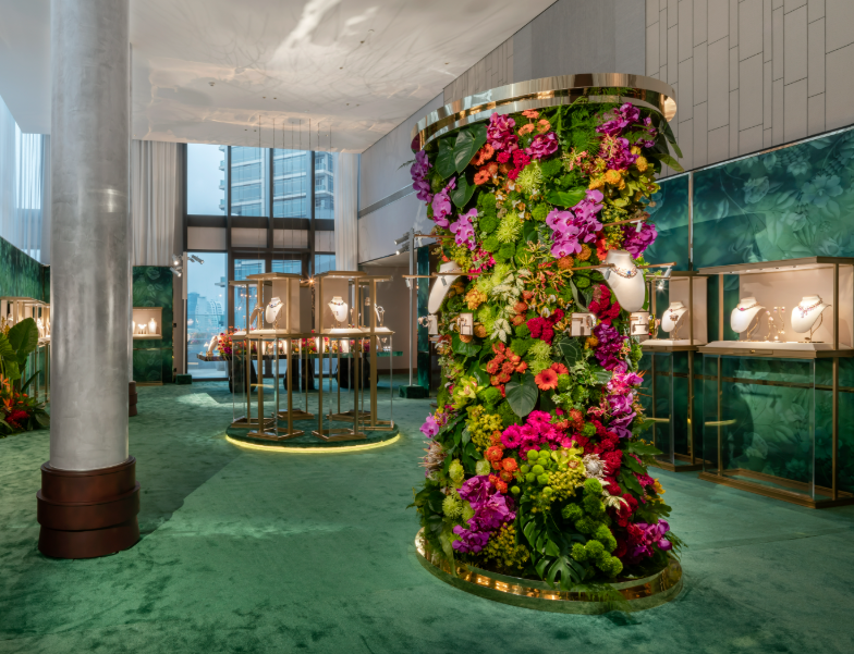 A high jewelry exhibition with a towering floral column of orchids and roses, surrounded by glass vitrines on emerald carpet