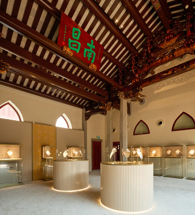 A high-jewelry exhibition hall with glass vitrines and tall cream pedestals beneath a dark timber-raftered ceiling