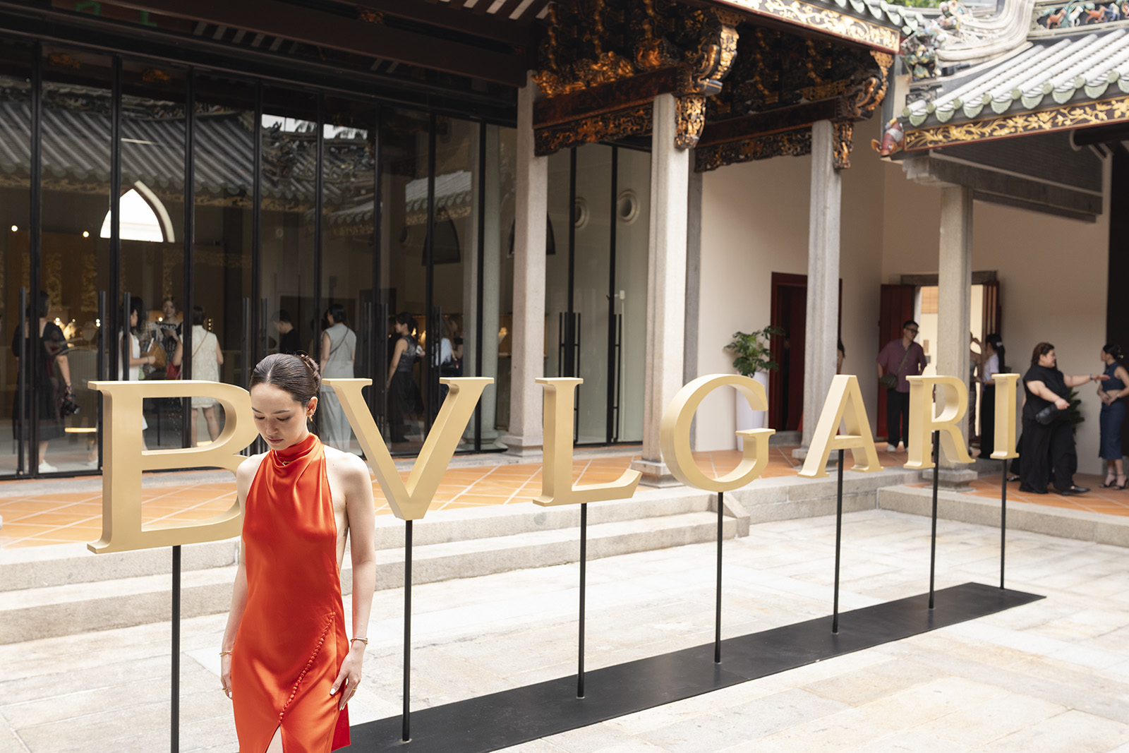 A guest in an orange gown standing beside large gold BVLGARI letters in the courtyard of House of Tan Yeok Nee